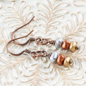 May include: A pair of handcrafted copper earrings with a hook design. Each earring features a series of beads in silver, copper, and gold colors. The earrings are displayed on a white, textured surface.