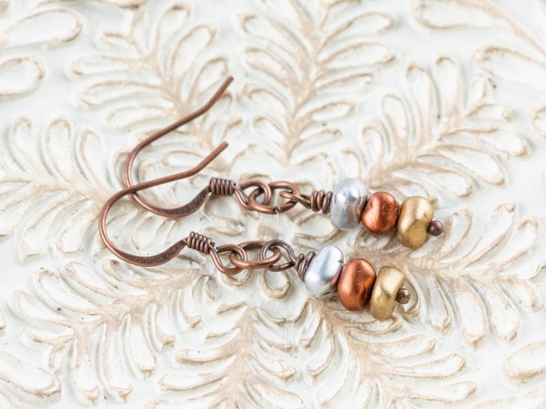 May include: A pair of handcrafted copper earrings with a hook design. Each earring features a series of beads in silver, copper, and gold colors. The earrings are displayed on a white, textured surface.