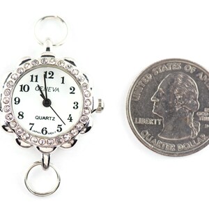 May include: A silver-toned Geneva quartz watch with a white face and a decorative, crystal-studded bezel. The watch has a ring at the top and bottom for attachment. A US quarter is next to the watch for size comparison.