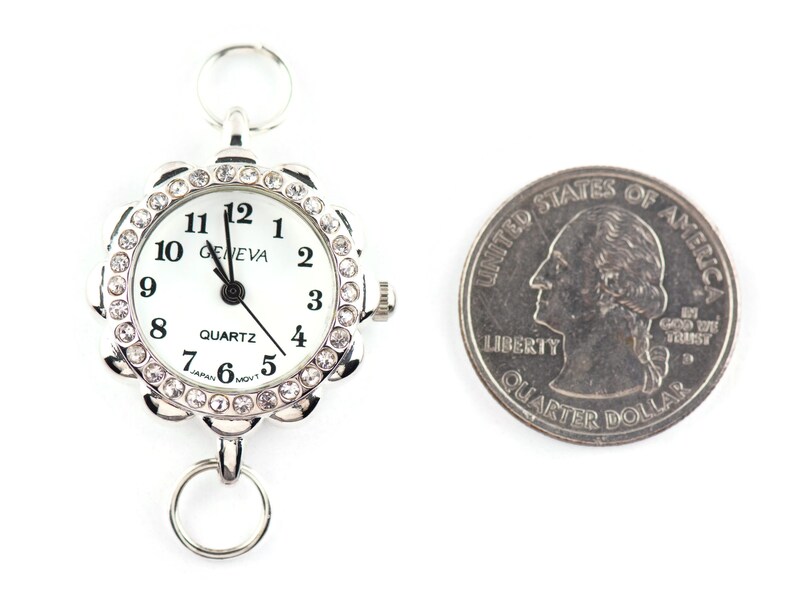 May include: A silver-toned Geneva quartz watch with a white face and a decorative, crystal-studded bezel. The watch has a ring at the top and bottom for attachment. A US quarter is next to the watch for size comparison.