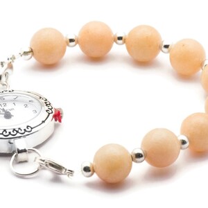 Puede incluir: Un reloj plateado con una esfera blanca y n&uacute;meros negros est&aacute; unido a una pulsera hecha de cuentas de color naranja claro.