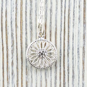 K&ouml;nnte beinhalten: Silberkette mit einem zarten, runden Anh&auml;nger mit filigranem Design. Der Anh&auml;nger hat ein zentrales blumenartiges Motiv mit filigranen Details.