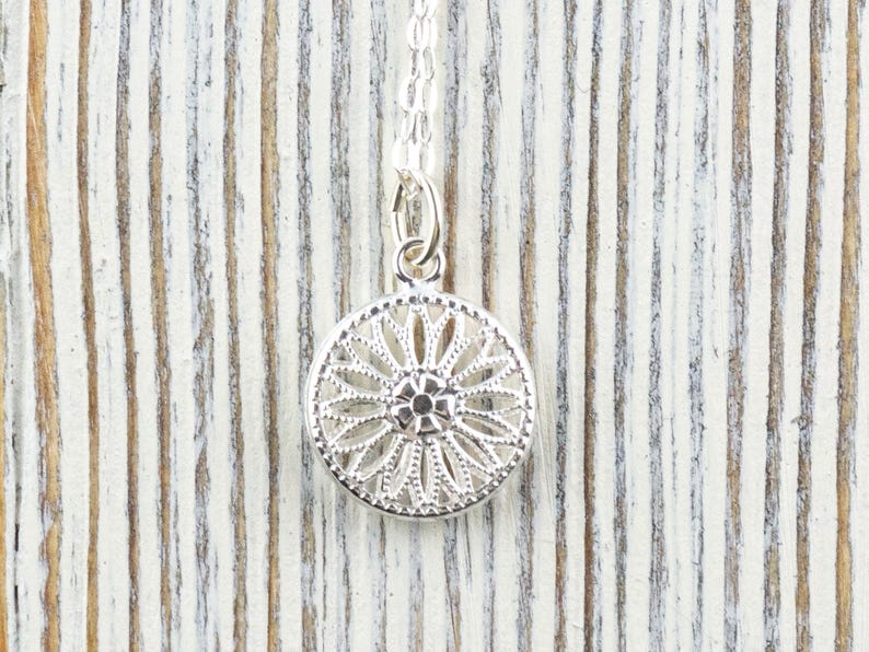 K&ouml;nnte beinhalten: Silberkette mit einem zarten, runden Anh&auml;nger mit filigranem Design. Der Anh&auml;nger hat ein zentrales blumenartiges Motiv mit filigranen Details.