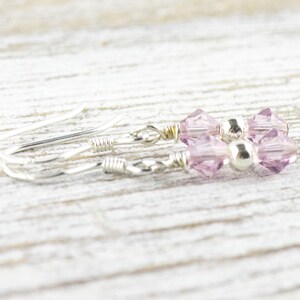 May include: A pair of delicate earrings featuring light purple crystal beads and a small silver bead. The earrings have silver-colored ear wires and are displayed on a light-colored wooden surface.