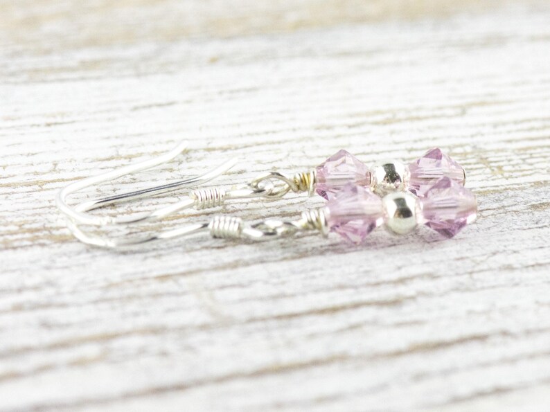 May include: A pair of delicate earrings featuring light purple crystal beads and a small silver bead. The earrings have silver-colored ear wires and are displayed on a light-colored wooden surface.