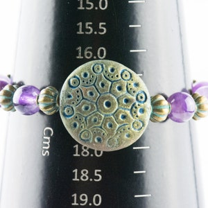 May include: A purple and gold beaded bracelet with a large, round, textured, green and gold charm. The bracelet is 18 cm in diameter.