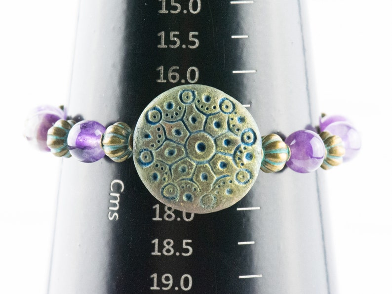 May include: A purple and gold beaded bracelet with a large, round, textured, green and gold charm. The bracelet is 18 cm in diameter.