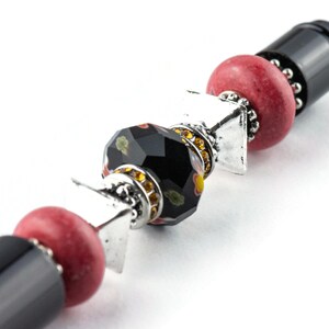 May include: A black and red beaded pen with silver accents. The pen has a black barrel with a red bead, a black bead with gold and red accents, a silver spacer, and a red bead.