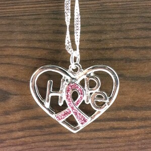Pode incluir: Colar com pingente de cora&ccedil;&atilde;o prateado com a palavra "HOPE" recortada. Uma fita rosa com detalhes brilhantes est&aacute; no centro do cora&ccedil;&atilde;o. O colar pende de uma corrente prateada.