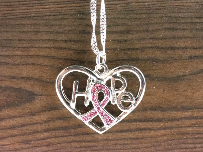 Pode incluir: Colar com pingente de cora&ccedil;&atilde;o prateado com a palavra "HOPE" recortada. Uma fita rosa com detalhes brilhantes est&aacute; no centro do cora&ccedil;&atilde;o. O colar pende de uma corrente prateada.