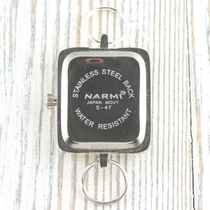 Puede incluir: Un reloj de acero inoxidable con una esfera cuadrada y un llavero plateado. La esfera del reloj es negra con texto blanco que dice "NARMI JAPAN MOVT S-47 WATER RESISTANT". El reloj est&aacute; sujeto a un llavero plateado.