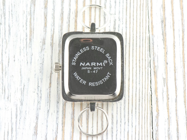 Puede incluir: Un reloj de acero inoxidable con una esfera cuadrada y un llavero plateado. La esfera del reloj es negra con texto blanco que dice "NARMI JAPAN MOVT S-47 WATER RESISTANT". El reloj est&aacute; sujeto a un llavero plateado.