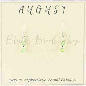 May include: A pair of silver earrings with green crystal beads. The earrings are displayed on a card with the text "AUGUST" and "Black Brook Shop".