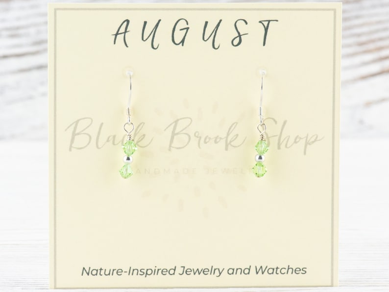 May include: A pair of silver earrings with green crystal beads. The earrings are displayed on a card with the text "AUGUST" and "Black Brook Shop".