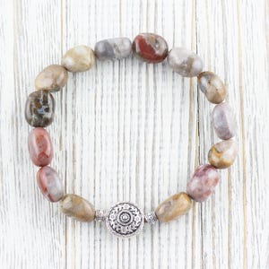 Op de afbeelding: Een armband gemaakt van bruine, grijze en roze stenen met een zilveren sluiting. De armband ligt op een witte houten achtergrond.