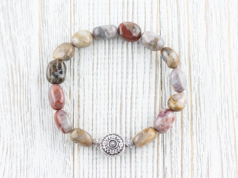 Op de afbeelding: Een armband gemaakt van bruine, grijze en roze stenen met een zilveren sluiting. De armband ligt op een witte houten achtergrond.