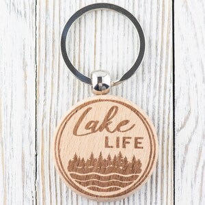 K&ouml;nnte beinhalten: Ein runder Holzschl&uuml;sselanh&auml;nger mit einem silbernen Schl&uuml;sselring. Der Schl&uuml;sselanh&auml;nger zeigt die Worte "Lake LIFE" in einem kreisf&ouml;rmigen Design, mit einer Wald- und Wasserszene darunter. Das Holz hat eine nat&uuml;rliche, hellbraune Farbe.