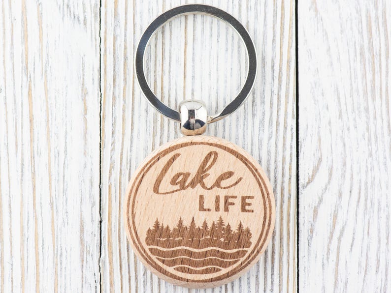 K&ouml;nnte beinhalten: Ein runder Holzschl&uuml;sselanh&auml;nger mit einem silbernen Schl&uuml;sselring. Der Schl&uuml;sselanh&auml;nger zeigt die Worte "Lake LIFE" in einem kreisf&ouml;rmigen Design, mit einer Wald- und Wasserszene darunter. Das Holz hat eine nat&uuml;rliche, hellbraune Farbe.
