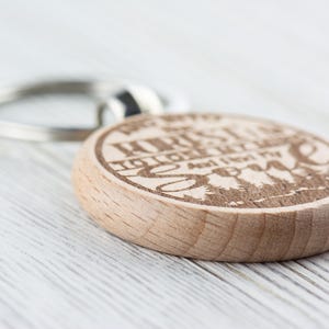 Op de afbeelding: Houten sleutelhanger met een rond ontwerp met de tekst "We're all just a little lost and looking for someone" en een silhouet van bomen. De sleutelhanger heeft een zilveren metalen ring.