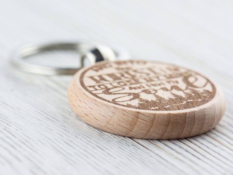 Op de afbeelding: Houten sleutelhanger met een rond ontwerp met de tekst "We're all just a little lost and looking for someone" en een silhouet van bomen. De sleutelhanger heeft een zilveren metalen ring.