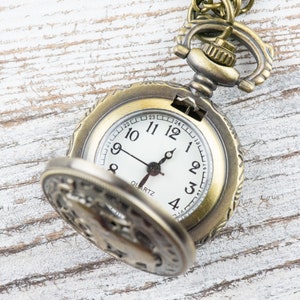May include: A gold-toned pocket watch with an open face, revealing a white clock face with black hands and black Roman numeral markings. The watch has a decorative design around the edge and a chain attached to the top.