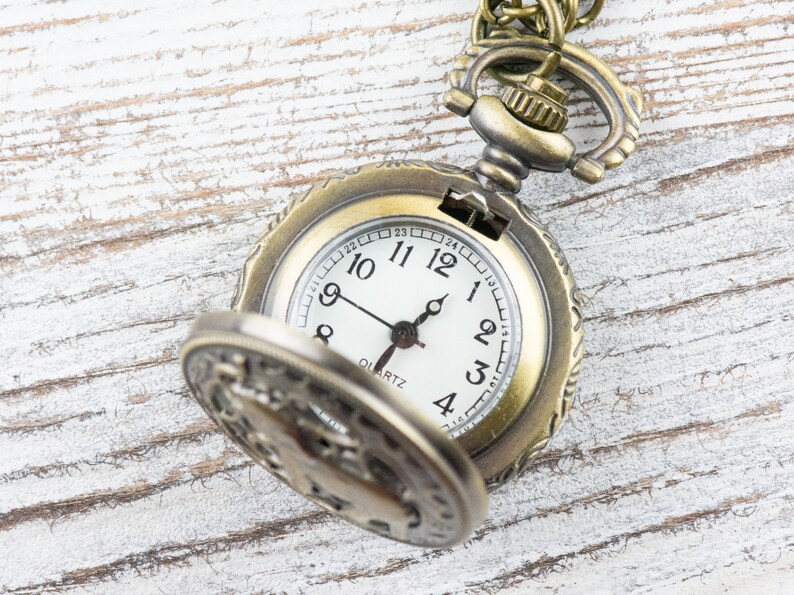 May include: A gold-toned pocket watch with an open face, revealing a white clock face with black hands and black Roman numeral markings. The watch has a decorative design around the edge and a chain attached to the top.
