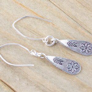 Puede incluir: Un par de pendientes de plata con forma de l&aacute;grima. Cada pendiente tiene un peque&ntilde;o dise&ntilde;o de flor y flecha.