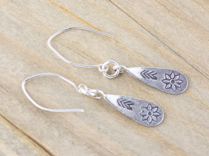 Puede incluir: Un par de pendientes de plata con forma de l&aacute;grima. Cada pendiente tiene un peque&ntilde;o dise&ntilde;o de flor y flecha.