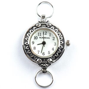 Op de afbeelding: Een zilverkleurig zakhorloge met een witte wijzerplaat en zwarte wijzers. Het horloge heeft een decoratieve lunette en twee ringen voor bevestiging. De wijzerplaat toont de merknaam "NARMI".
