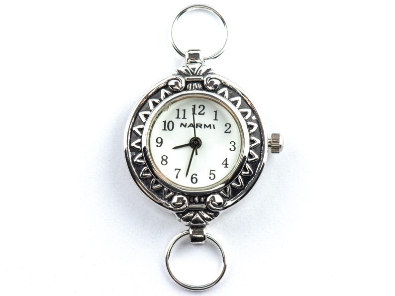 Op de afbeelding: Een zilverkleurig zakhorloge met een witte wijzerplaat en zwarte wijzers. Het horloge heeft een decoratieve lunette en twee ringen voor bevestiging. De wijzerplaat toont de merknaam "NARMI".