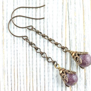May include: A pair of drop earrings with a bronze chain design. Each earring features a purple gemstone, encased in a decorative bronze cap. The earrings are presented on a white wooden surface. The gemstones are approximately 1 cm in diameter.