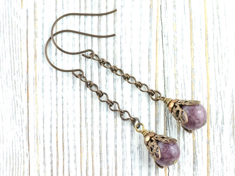 May include: A pair of drop earrings with a bronze chain design. Each earring features a purple gemstone, encased in a decorative bronze cap. The earrings are presented on a white wooden surface. The gemstones are approximately 1 cm in diameter.