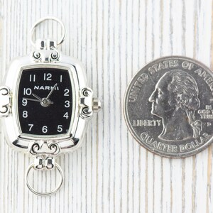 Pu&ograve; includere: Orologio da tasca argentato con quadrante nero e numeri bianchi. L'orologio ha una forma rettangolare con bordi arrotondati e un anello nella parte superiore per attaccare una catena. Una moneta da 25 centesimi americana &egrave; accanto all'orologio per il confronto delle dimensioni.