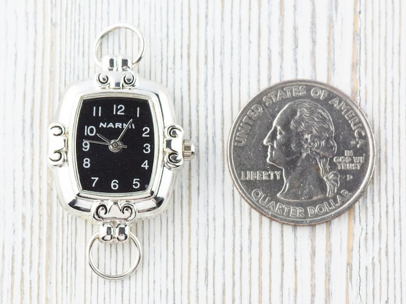 Pu&ograve; includere: Orologio da tasca argentato con quadrante nero e numeri bianchi. L'orologio ha una forma rettangolare con bordi arrotondati e un anello nella parte superiore per attaccare una catena. Una moneta da 25 centesimi americana &egrave; accanto all'orologio per il confronto delle dimensioni.