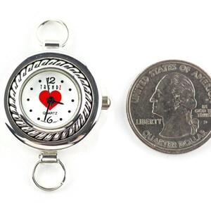 May include: Silver quartz watch with a white face, red heart, and the word "TRENDZ". The watch has a braided bezel and attachment loops. A U.S. quarter is next to the watch for size comparison. The watch is approximately 3.8 cm in diameter.