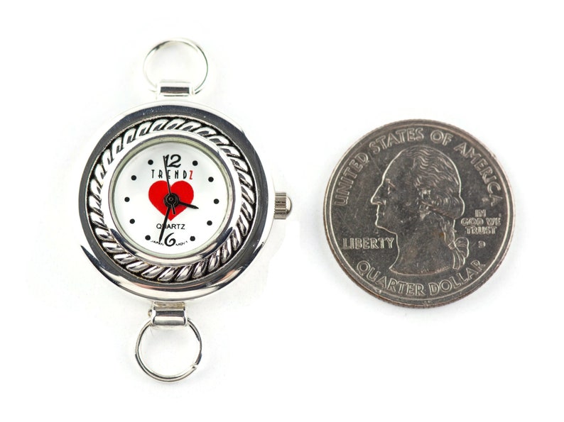 May include: Silver quartz watch with a white face, red heart, and the word "TRENDZ". The watch has a braided bezel and attachment loops. A U.S. quarter is next to the watch for size comparison. The watch is approximately 3.8 cm in diameter.