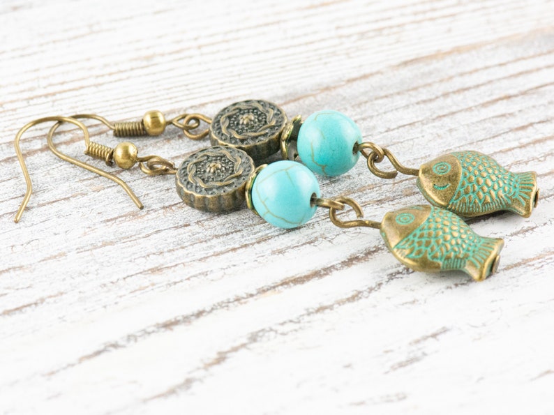 May include: A pair of dangle earrings with turquoise beads and bronze fish charms. The earrings have a floral design on the top of the beads.