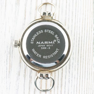 May include: The back of a silver pocket watch with the text "Stainless Steel Back", "Narmi", "Japan Movt SNW - 8", and "Water Resistant".