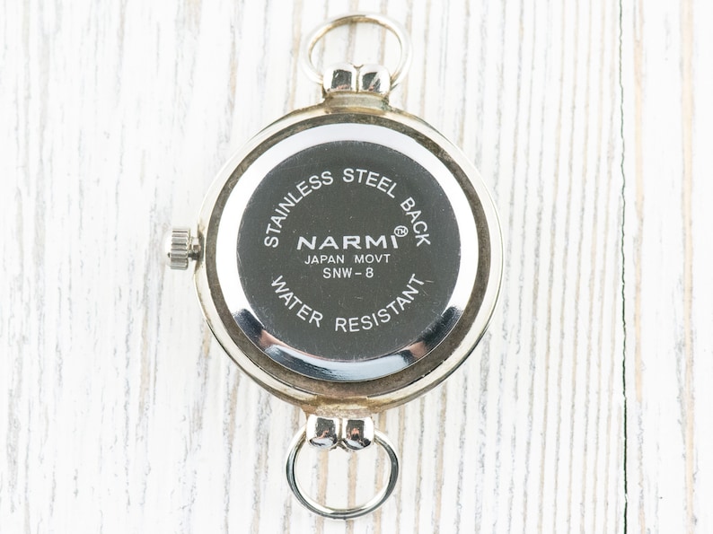 May include: The back of a silver pocket watch with the text "Stainless Steel Back", "Narmi", "Japan Movt SNW - 8", and "Water Resistant".
