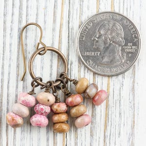Op de afbeelding: Handgemaakte oorbellen met een bronzen metalen haak en ring. De oorbellen zijn versierd met kleine, kleurrijke, ovaalvormige kralen in tinten roze, beige en bruin. Een munt is inbegrepen ter referentie van de grootte.