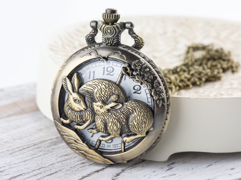 Puede incluir: Reloj de bolsillo de bronce antiguo con un dise&ntilde;o detallado de conejos. La esfera del reloj es blanca con n&uacute;meros y manecillas negras. Se adjunta una cadena a juego. Un accesorio cl&aacute;sico.