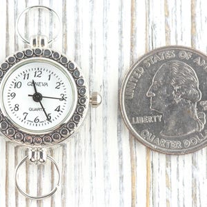 May include: Silver-toned Geneva quartz watch with a white face, black numerals, and a decorative bezel. The watch is next to a US quarter for size comparison. The watch has a ring at the top and bottom for attachment.