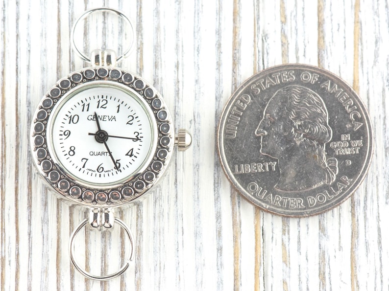 May include: Silver-toned Geneva quartz watch with a white face, black numerals, and a decorative bezel. The watch is next to a US quarter for size comparison. The watch has a ring at the top and bottom for attachment.
