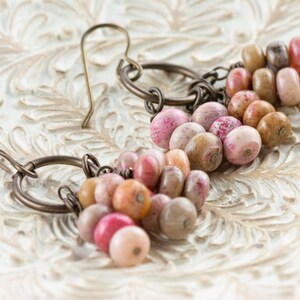 Op de afbeelding: Handgemaakte oorbellen met een reeks kleurrijke kralen in roze, perzik en bruin. De kralen zijn geregen en bevestigd aan bronzen metalen ringen en oorhaken. De oorbellen liggen op een wit, sierlijk oppervlak.