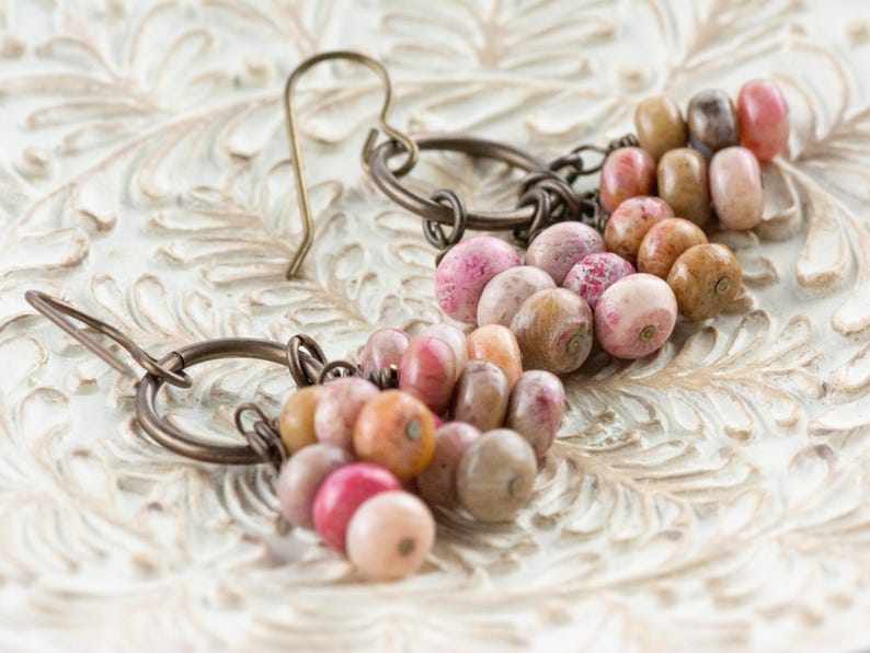 Op de afbeelding: Handgemaakte oorbellen met een reeks kleurrijke kralen in roze, perzik en bruin. De kralen zijn geregen en bevestigd aan bronzen metalen ringen en oorhaken. De oorbellen liggen op een wit, sierlijk oppervlak.