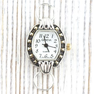 Peut inclure: Cadran de montre Geneva de forme ovale avec un cadran blanc, des aiguilles noires et des d&eacute;tails dor&eacute;s. La montre a un cadre argent&eacute; avec des clous dor&eacute;s et une couronne dor&eacute;e. Elle est fix&eacute;e &agrave; des boucles argent&eacute;es.