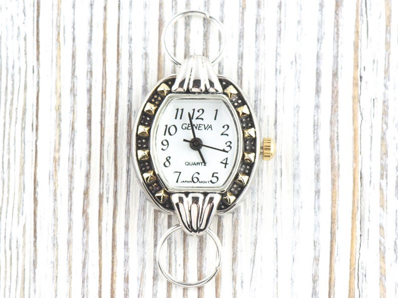 Peut inclure: Cadran de montre Geneva de forme ovale avec un cadran blanc, des aiguilles noires et des d&eacute;tails dor&eacute;s. La montre a un cadre argent&eacute; avec des clous dor&eacute;s et une couronne dor&eacute;e. Elle est fix&eacute;e &agrave; des boucles argent&eacute;es.
