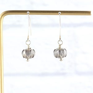Pu&ograve; includere: Un paio di orecchini pendenti in argento. Ogni orecchino presenta una perla rotonda e dettagliata in argento con un motivo a forma di foglia. Gli orecchini pendono da un semplice gancio d'argento. Gli orecchini sono esposti su un supporto dorato.