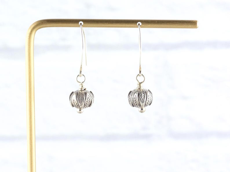 Pu&ograve; includere: Un paio di orecchini pendenti in argento. Ogni orecchino presenta una perla rotonda e dettagliata in argento con un motivo a forma di foglia. Gli orecchini pendono da un semplice gancio d'argento. Gli orecchini sono esposti su un supporto dorato.