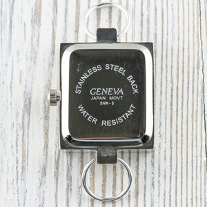 K&ouml;nnte beinhalten: Silberne quadratische Uhr mit einem Edelstahlboden. Das Zifferblatt der Uhr zeigt "Geneva Japan Movt SNW-5 Water Resistant".